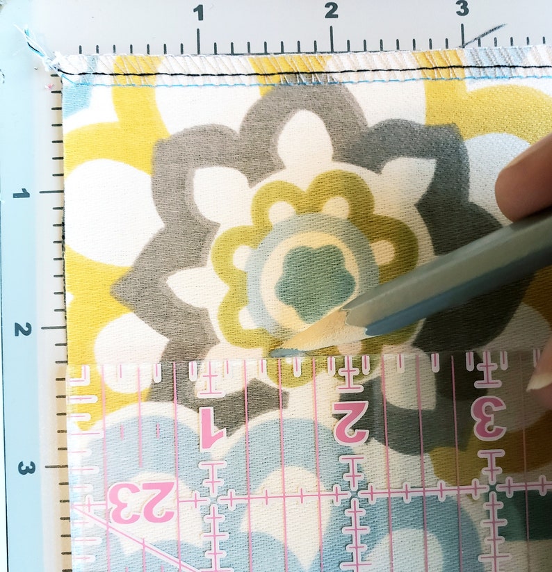 May include: A close-up of a fabric with a floral pattern in shades of yellow, gray, and blue. A ruler with markings in inches is visible, and a pencil is being used to mark a point on the fabric.