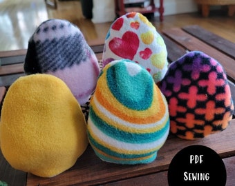 May include: Five colorful fleece egg-shaped toys with different patterns. The toys are arranged in a circle on a wooden surface. The text "PDF SEWING PATTERN" is visible in the lower right corner of the image.
