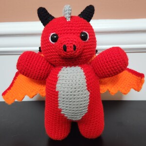 May include: A red crocheted dragon plush toy with orange wings. The dragon has black horns and large black eyes.