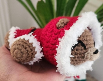 May include: A handmade crochet bear wearing a red Santa suit with white trim. The bear has brown fur, a black stitched nose and closed eyes. The bear is lying down, and the Santa suit has a brown button.