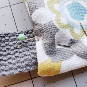 May include: Close-up of two fabric pieces pinned together with a sewing pin. The fabric on the left is a gray textured material. The fabric on the right is a white fabric with a gray, yellow, and blue floral pattern.