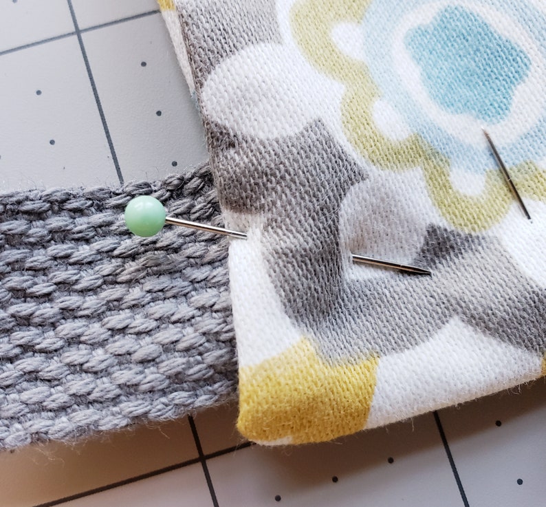 May include: Close-up of two fabric pieces pinned together with a sewing pin. The fabric on the left is a gray textured material. The fabric on the right is a white fabric with a gray, yellow, and blue floral pattern.