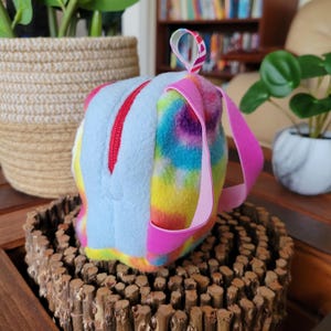 May include: A small, blue and rainbow tie-dye fleece backpack with pink straps. The backpack has a zipper closure and a small loop on the top.