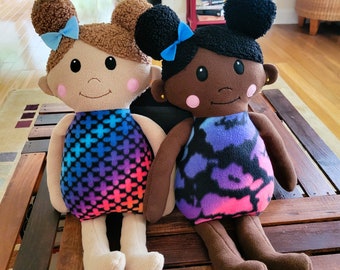 May include: Two handmade fabric dolls with brown and black hair, wearing colorful patterned dresses. The doll on the left has light skin and blue eyes, while the doll on the right has dark skin and brown eyes.