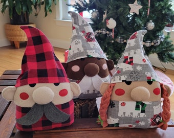 May include: Three Christmas gnome decorations with fabric hats and plush faces. One gnome has a red and black plaid hat and a gray mustache. The other two gnomes have gray hats with polar bear patterns. One gnome has a white beard and the other has braided hair.