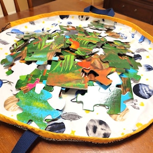 以下が含まれることがあります： 青いハンドルが付いた白と黄色の布製の収納袋。袋には、ジャングルのシーンが描かれたカラフルなパズルが入っています。