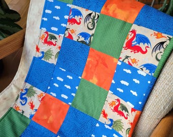 May include: A patchwork quilt featuring squares of blue, green, orange, and beige fabric. The quilt is decorated with dragon motifs in red, blue, and green, with flames and clouds. The quilt is folded over a chair.