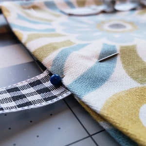 May include: Close-up of a fabric with a blue, yellow, and white geometric pattern. A black and white checkered ribbon is pinned to the fabric with a blue pin.