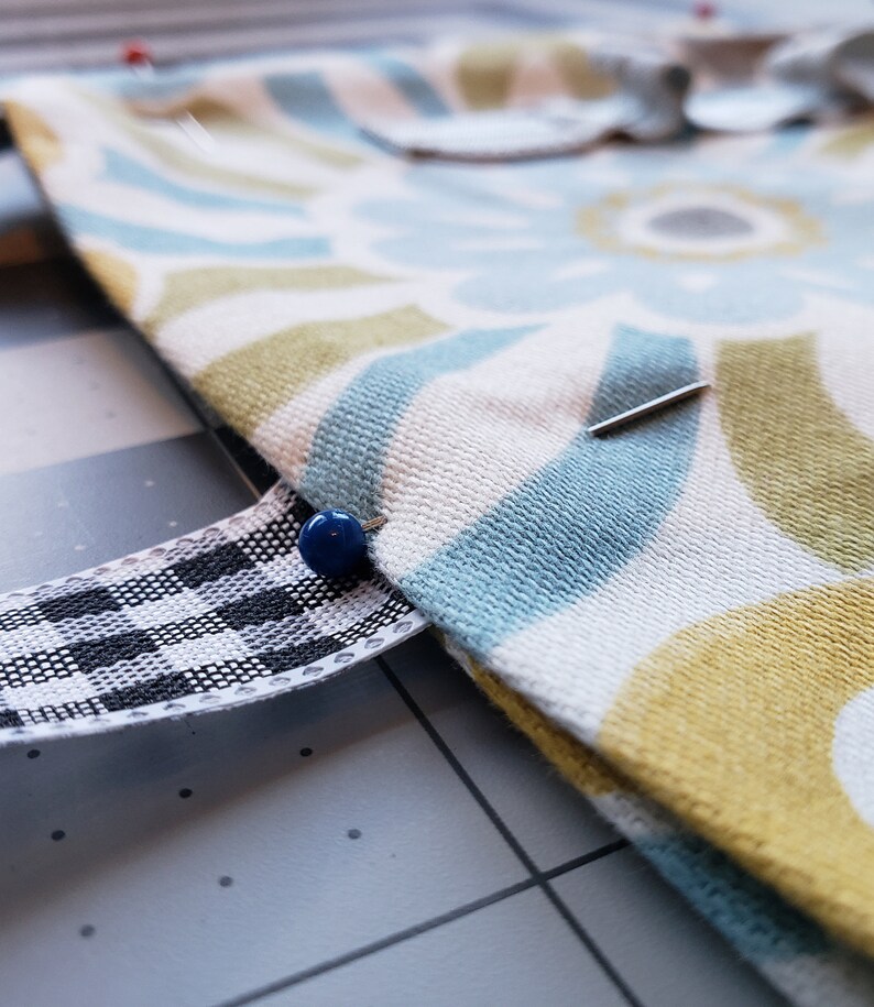 May include: Close-up of a fabric with a blue, yellow, and white geometric pattern. A black and white checkered ribbon is pinned to the fabric with a blue pin.
