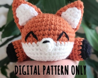 May include: A crocheted fox amigurumi toy with orange fur, white facial features, and black accents on the paws. The toy is being held in a hand, and the text "DIGITAL PATTERN ONLY" is visible below the toy.