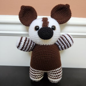 May include: A crocheted stuffed animal toy in the shape of a dog. The toy is brown and white with black eyes and a black nose. The toy is standing on a black surface.