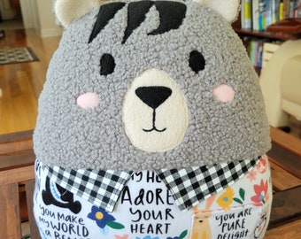 May include: A gray plush bear with black eyes and a black nose. The bear is wearing a black and white checkered collar and a white shirt with a floral pattern. The shirt has the text "You make my world a better place" and "I love you" on it. The bear is sitting on a wooden surface.
