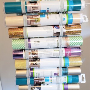 May include: A fabric tote bag with a floral pattern and black and white checkered straps, filled with rolls of vinyl in various colors, including teal, gold, silver, and black. The vinyl rolls are labeled "Cricut" and have different finishes, such as glitter, permanent, and removable.