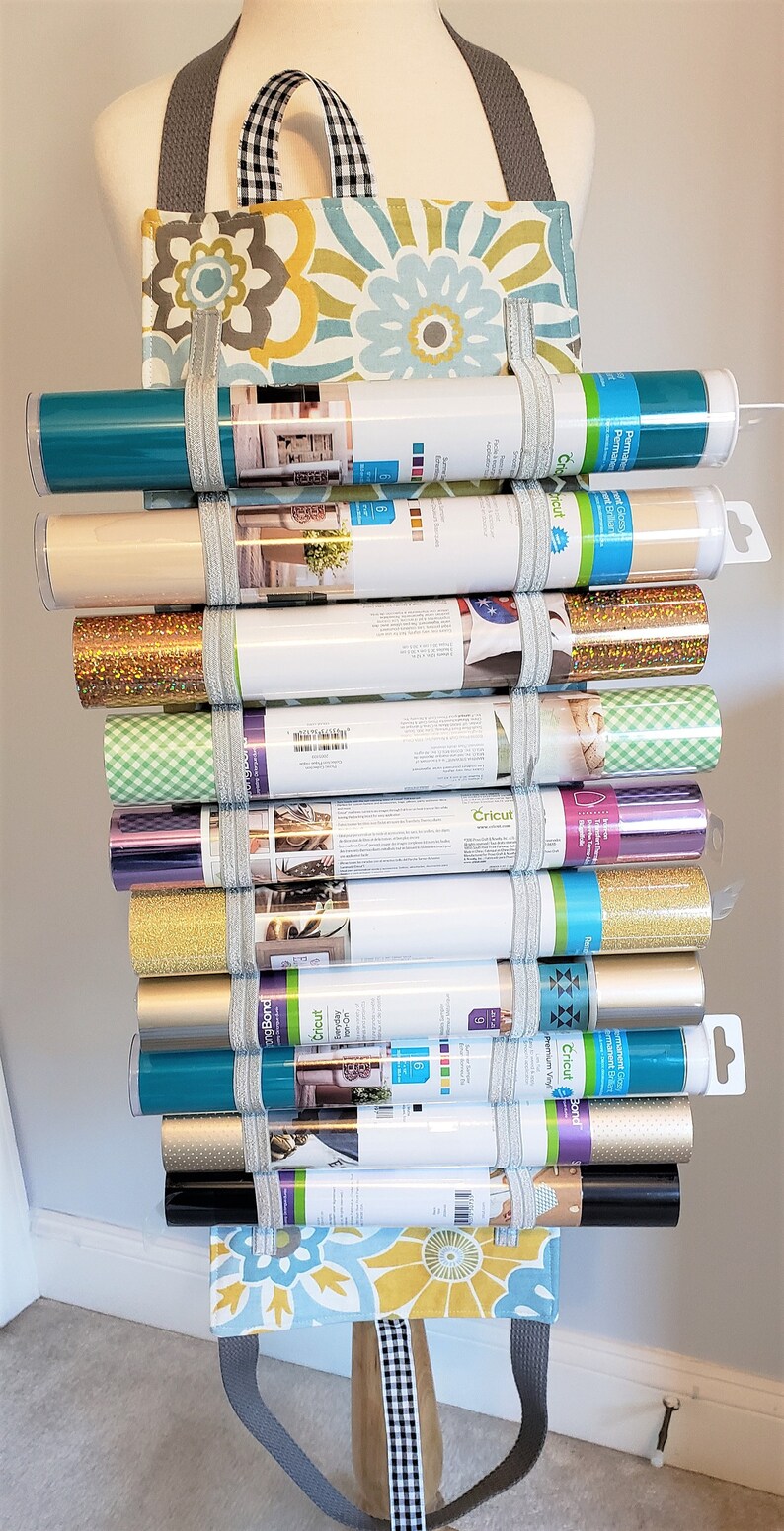 May include: A fabric tote bag with a floral pattern and black and white checkered straps, filled with rolls of vinyl in various colors, including teal, gold, silver, and black. The vinyl rolls are labeled "Cricut" and have different finishes, such as glitter, permanent, and removable.