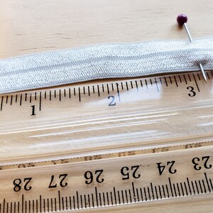 May include: A silver elastic band measuring 2 inches in length, pinned to a ruler with a purple pin.