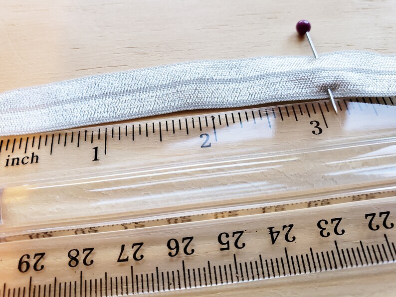 May include: A silver elastic band measuring 2 inches in length, pinned to a ruler with a purple pin.