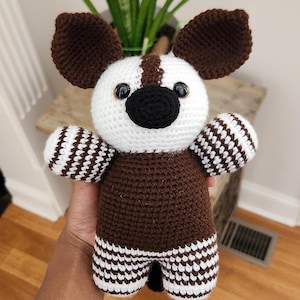 May include: A handmade crochet okapi plush toy featuring a white head with a dark brown stripe, large dark brown ears, and a black nose. The body is dark brown with black and white striped legs and arms, and two shiny black plastic safety eyes.