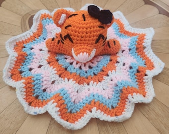 May include: A crochet security blanket featuring an orange tiger head with black stripes and a small black bow. The blanket base is a circular, multi-colored star-shaped design with concentric rings of white, light blue, pink, and orange yarn.