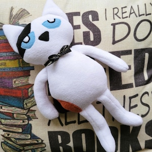 May include: White plush cat toy with black markings and blue eyes. The cat is wearing a black bow and has a brown patch on its side. The cat is lying on a beige pillow with the text "I really do love all the books" printed on it.