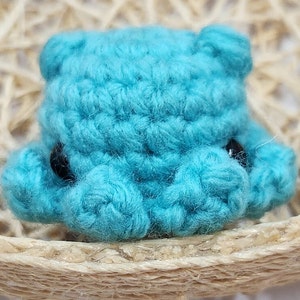 May include: A small crocheted blue bear with black eyes. The bear is sitting on a woven straw surface.