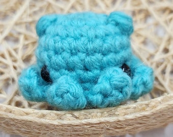 May include: A small crocheted blue bear with black eyes. The bear is sitting on a woven straw surface.
