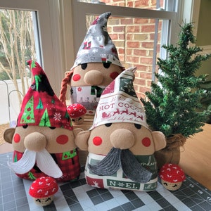 May include: Three handmade Christmas gnome decorations. The gnomes are made of felt and fabric and have different patterns. The gnomes are wearing hats and have beards. The gnomes are sitting on a table with a small Christmas tree in the background.