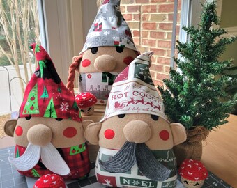 May include: Three handmade Christmas gnome ornaments. The gnomes are made of felt and fabric and have different patterns. The gnomes are wearing hats and have beards. The gnomes are sitting on a table with a small Christmas tree in the background.