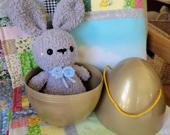 May include: A gray plush bunny with long ears and a blue bow tie sits inside a gold-colored plastic egg. The egg is open, with the top beside it. The bunny is on a colorful patchwork quilt.