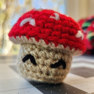 May include: A crocheted mushroom toy with a red cap and a tan body. The mushroom has a simple, smiling face.