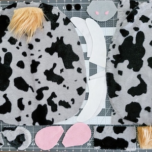 May include: A set of fabric pieces for a cow costume. The pieces are cut out of gray and black fabric with a cow print, white felt, pink felt, and brown faux fur. The pieces include the head, ears, horns, and tail.