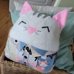 May include: A white plush cat pillow with pink ears and a gray stripe on its head. The pillow is wearing a blue and beige patchwork shirt with a pocket featuring a smaller cat illustration.
