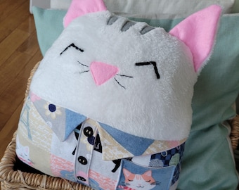 May include: A white plush cat pillow with pink ears and a gray stripe on its head. The pillow is wearing a blue and beige patchwork shirt with a pocket featuring a smaller cat illustration.