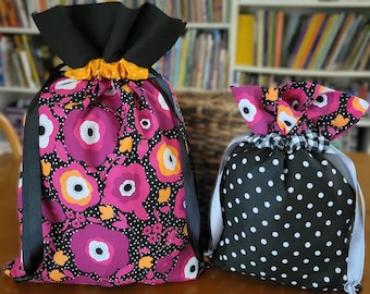 May include: Two fabric drawstring bags. The larger bag is black with pink and yellow floral print. The smaller bag is black with white polka dots.