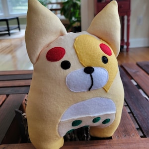 Op de afbeelding: Een pluche speelgoed in de vorm van een hond met een geel, wit en rood ontwerp. Het speelgoed heeft een witte neus en groene vlekken op zijn buik.