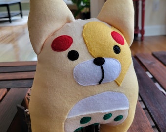 May include: A plush toy shaped like a dog with a yellow, white, and red design. The toy has a white nose and green spots on its belly.