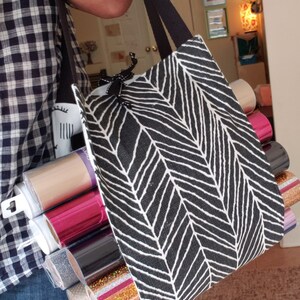 May include: A black and white tote bag with a chevron pattern, filled with rolls of colorful vinyl. The bag has black straps and a black zipper.