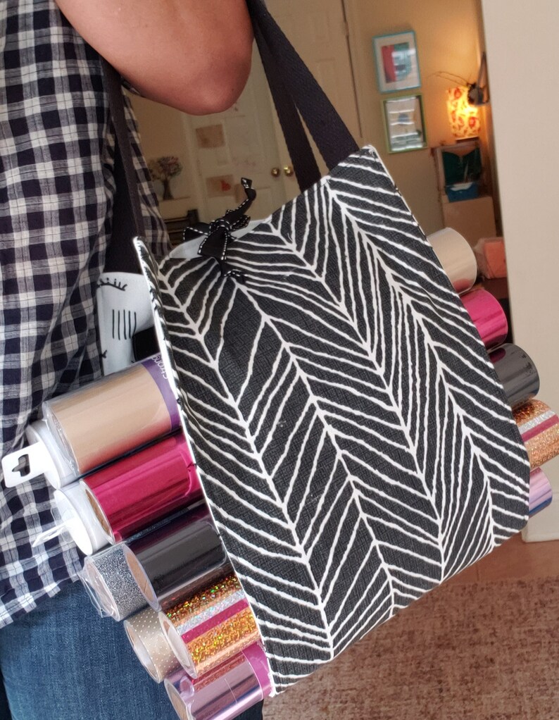 May include: A black and white tote bag with a chevron pattern, filled with rolls of colorful vinyl. The bag has black straps and a black zipper.
