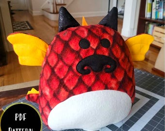 May include: A red and yellow plush dragon toy with black eyes and a black nose. The dragon is sitting on a gray surface with a ruler and the words "PDF PATTERN" in a yellow circle.