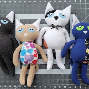 May include: Four plush cat toys with different colors and patterns. The first cat is black with green eyes, the second is tan with a pink and purple heart pattern, the third is white with black spots and blue eyes, and the fourth is blue with green eyes and a yellow and black pattern. The blue cat has the text "Let's Go!" on its belly.