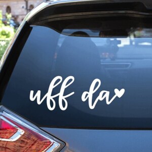 May include: A white vinyl sticker with the text "uff da" and a small heart on a grey car window.
