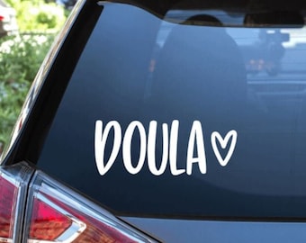 May include: A white vinyl sticker with the word "DOULA" and a small heart on a dark gray car window.