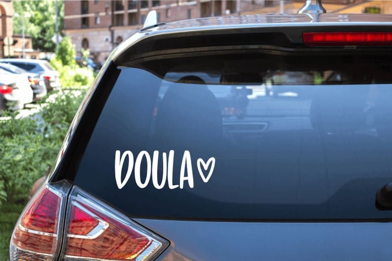 May include: A white vinyl sticker with the word "DOULA" and a small heart on a dark gray car window.