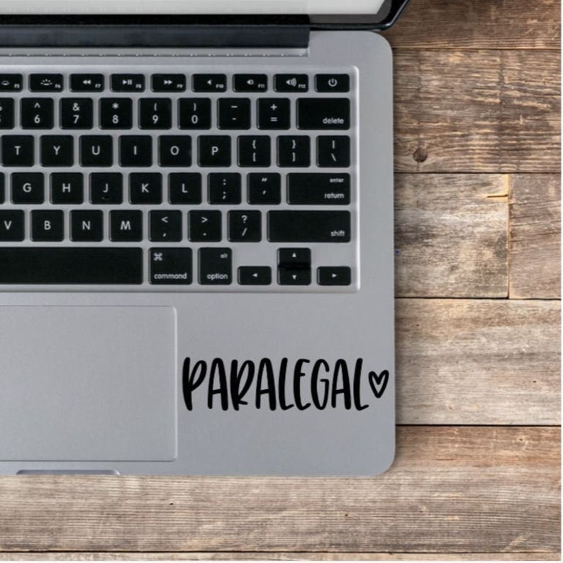 May include: A silver laptop computer with a black keyboard and a black trackpad. The trackpad has a black vinyl sticker with the text "PARALEGAL" and a small heart.