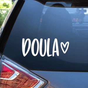 May include: A white vinyl sticker with the word "DOULA" and a small heart. The sticker is on the back window of a silver car.