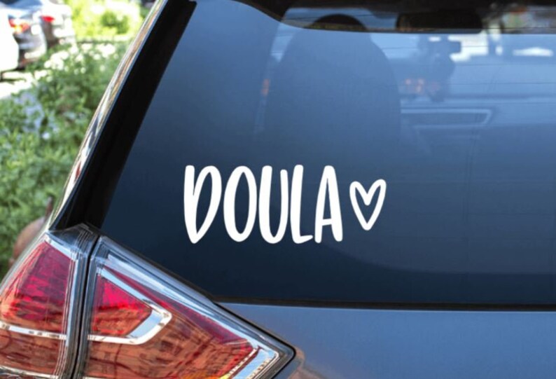 May include: A white vinyl sticker with the word "DOULA" and a small heart. The sticker is on the back window of a silver car.