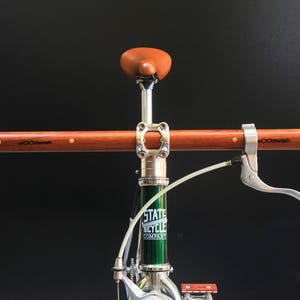 May include: A green bicycle with a wooden handlebar, a brown leather saddle, and a silver brake lever. The bicycle frame is labeled "State Bicycle Company".