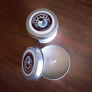 May include: Two silver-colored candle tins with lids, one open to reveal a white candle and wick. The lids feature a circular label with the text "shieldmaiden candleworks" and a graphic of a woman's profile.