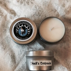 May include: Two round silver metal candle tins and one lid. One tin is open, showing white wax and a centered wick. The lid features a circular logo with the text Shieldmaiden candleworks and a silhouette. A side label reads Skadi's Embrace.