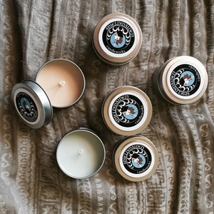 May include: Five round metal candle tins, some with lids on and some open to reveal light-colored wax and a central wick. Each lid features a circular black label with white moon phases, a blue profile illustration, and the text SHIELDMAIDEN candleworks.