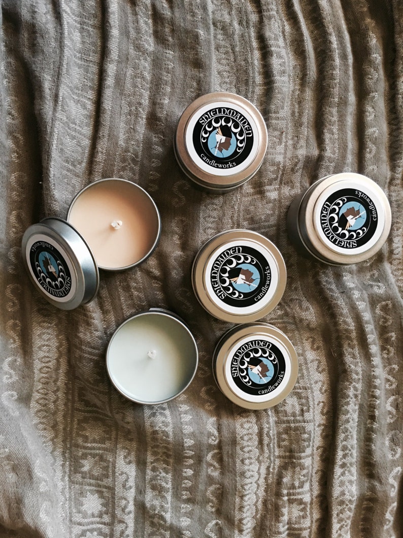 May include: Five round metal candle tins, some with lids on and some open to reveal light-colored wax and a central wick. Each lid features a circular black label with white moon phases, a blue profile illustration, and the text SHIELDMAIDEN candleworks.